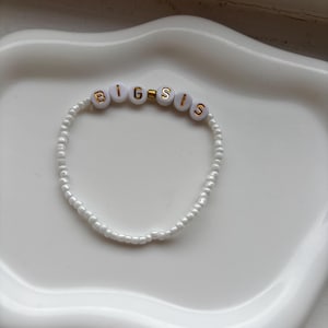 Op de afbeelding: Witte kralenarmband met de woorden "BIG SIS" in gouden en witte letterkralen. De armband wordt gepresenteerd op een wit, golvend dienblad.