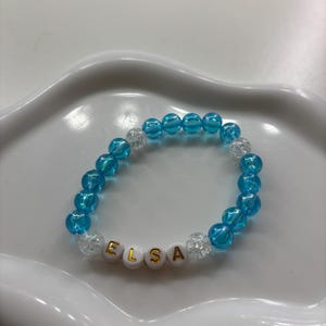 Könnte beinhalten: Ein Armband aus blauen und transparenten Perlen mit dem Namen "ELSA" in goldenen Buchstaben auf weißen Perlen. Das Armband wird auf einem weißen Tablett mit gewelltem Rand präsentiert.
