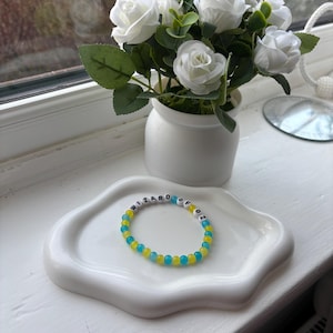 May include: A white ceramic cloud-shaped dish holds a beaded bracelet. The bracelet features yellow and blue beads spelling "WIZARD OF OZ". A small white vase with white roses sits in the background.