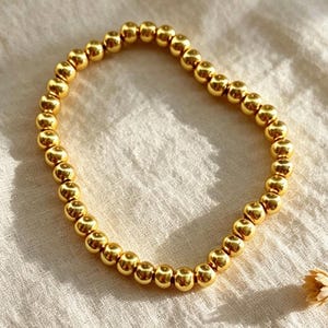 May include: A circular bracelet composed of numerous small, polished gold-toned spherical beads strung together. The beads have a reflective, metallic surface that catches the light, creating a uniform and continuous loop of golden spheres.