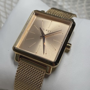 May include: A gold-coloured Nixon watch with a square face and mesh strap. The watch face is a light rose gold colour with gold hands and markers. The strap is a fine mesh of gold-coloured metal.