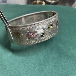 May include: Silver hinged bangle bracelet with floral and leaf design. The bracelet features embossed flowers with pink and gold accents. The bracelet is held by silver tongs against a green background.