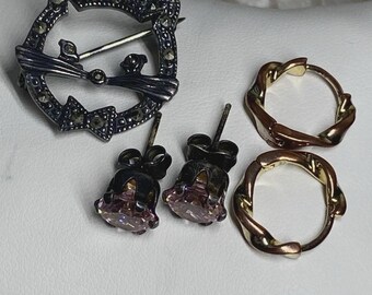 Puede incluir: Una colección de joyas que incluye un broche de gato plateado con detalles de marcasita, dos pendientes de botón con piedras rosas y dos pendientes de aro dorados. Las joyas se exhiben sobre una superficie blanca.