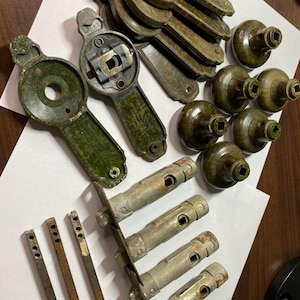 May include: An assortment of vintage door hardware, including olive green and brown doorknobs, backplates, and silver-toned latches. The collection also features several small, rectangular metal pieces, all arranged on a white surface.