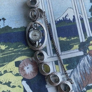 Könnte beinhalten: Silberfarbene Vintage-Uhr mit weißem Zifferblatt und dem Aufdruck "CARD". Die Uhr hat ein ovales Zifferblatt und eine Reihe von kreisförmigen Gliedern. Eine zarte Kette verbindet die Uhr mit dem Verschluss.