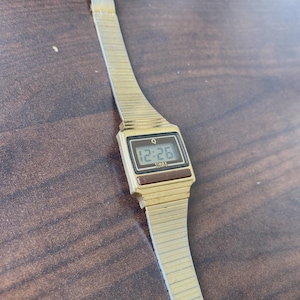 May include: A gold-coloured Timex Q digital watch showing the time 12:26. The watch has a rectangular face with a black digital display and a gold-coloured, ribbed metal strap. The watch is on a brown surface.