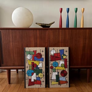 May include: Two rectangular wall art panels featuring a textured, multi-colored geometric mosaic design on a beige fabric background. The abstract composition includes circles, triangles, and rectangles in red, blue, green, and yellow, accented by black lines and starburst motifs.