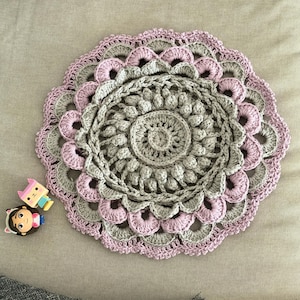 May include: A handmade, circular crochet doily in shades of gray and pink. The doily features a detailed, layered design with a scalloped edge. Decorative accents include two cartoon character magnets.