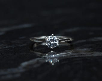 May include: A silver ring with a large, round, clear gemstone set in a six-prong setting. The ring is set against a dark, reflective surface, creating a mirrored effect of the ring.