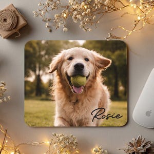 May include: A rectangular mouse pad featuring a photographic print of a golden retriever dog holding a yellow tennis ball in its mouth. The name Rosie is printed in a black cursive script in the bottom right corner of the mouse pad surface.