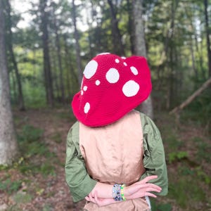May include: A red mushroom-shaped hat with white polka dots. The hat is crocheted and covers the head. The person is wearing a green shirt and khaki vest. The background is a forest with tall trees.