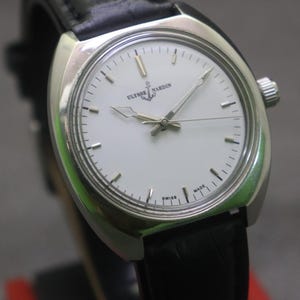 May include: A silver-toned Ulysse Nardin wristwatch with a white face and black leather band. The watch features a minimalist design with silver hands and hour markers. The watch face displays the brand name and the word "SWISS MADE".