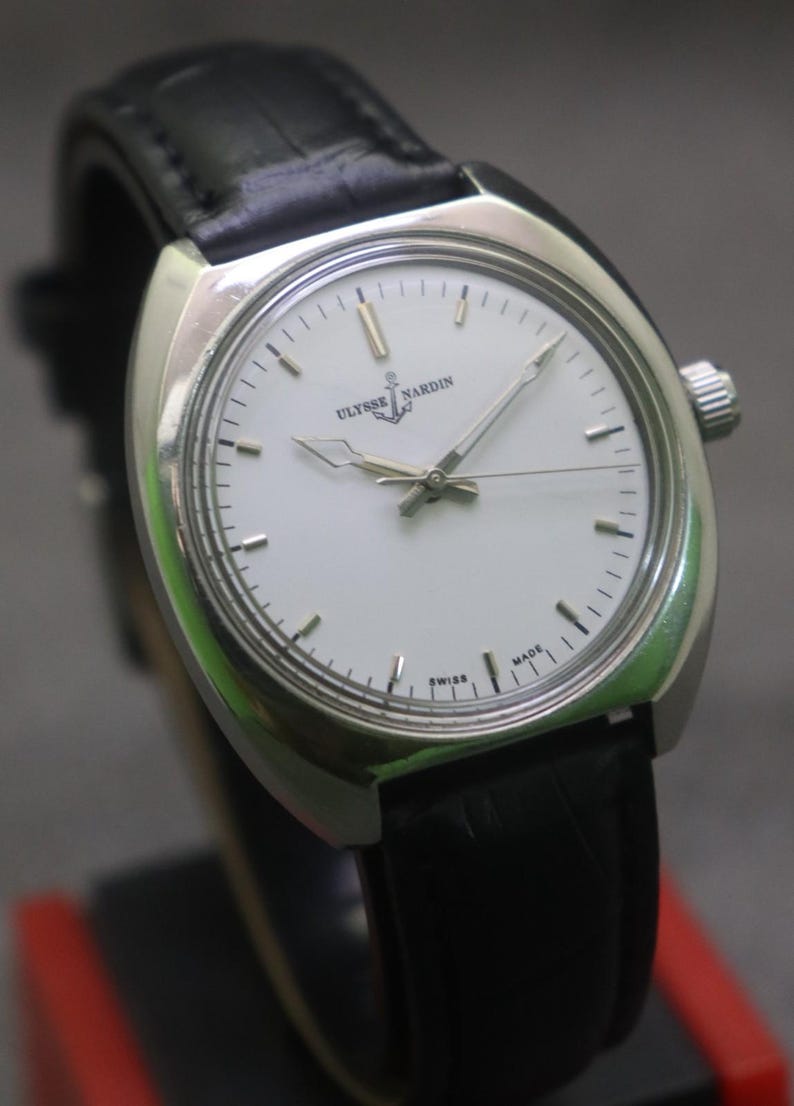 May include: A silver-toned Ulysse Nardin wristwatch with a white face and black leather band. The watch features a minimalist design with silver hands and hour markers. The watch face displays the brand name and the word "SWISS MADE".