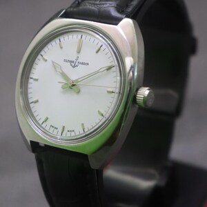 May include: A silver-toned Ulysse Nardin wristwatch with a white face and black leather band. The watch has a round face with silver hands and markers. The band is black and appears to be made of leather. The watch is displayed on a black stand.