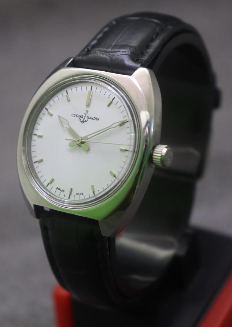May include: A silver-toned Ulysse Nardin wristwatch with a white face and black leather band. The watch has a round face with silver hands and markers. The band is black and appears to be made of leather. The watch is displayed on a black stand.