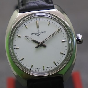 May include: A silver-toned Ulysse Nardin wristwatch with a white face and black leather band. The watch face features black hands and markers, with the brand name and anchor logo. The watch is marked "SWISS MADE".