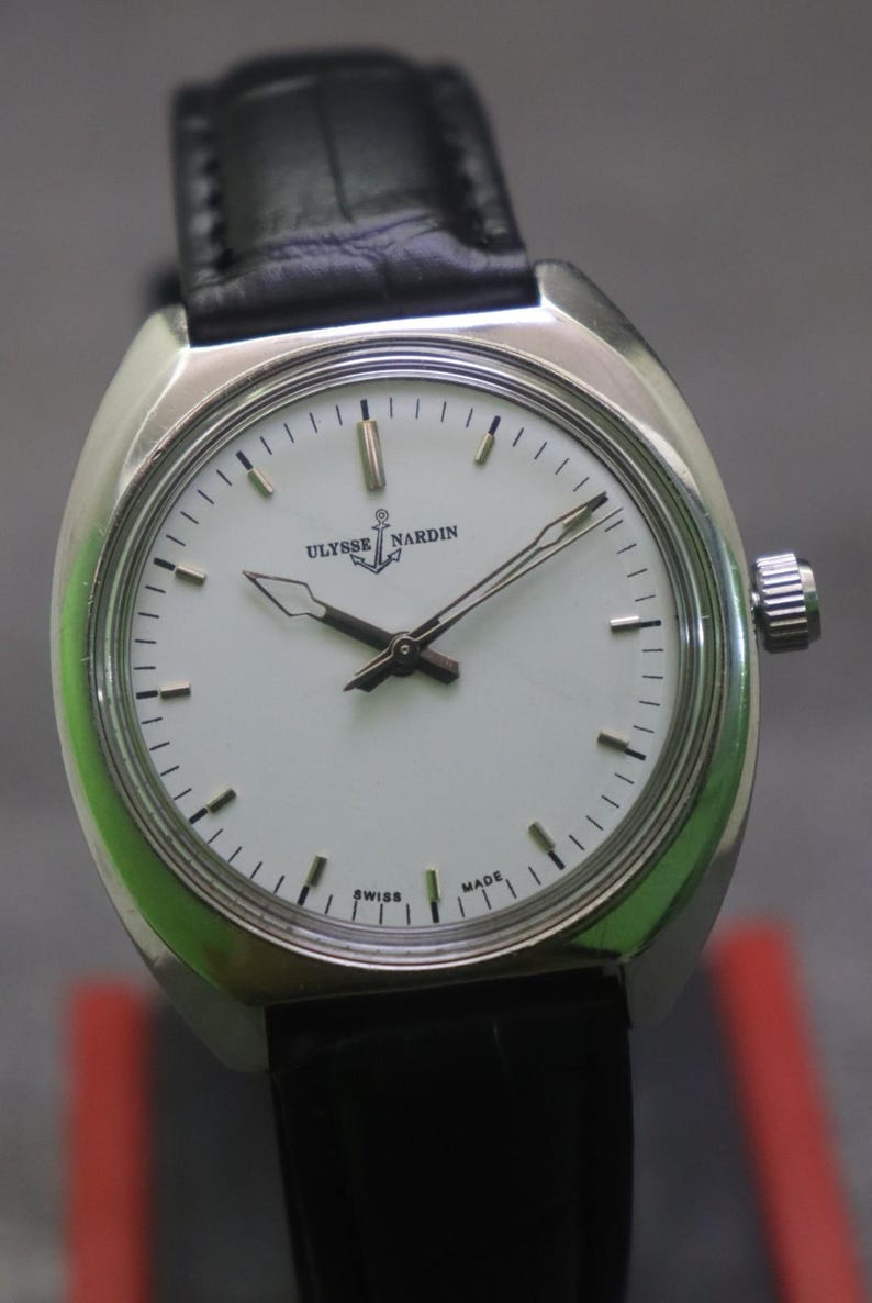 May include: A silver-toned Ulysse Nardin wristwatch with a white face and black leather band. The watch face features black hands and markers, with the brand name and anchor logo. The watch is marked "SWISS MADE".