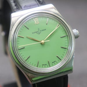 May include: A silver Ulysse Nardin wristwatch with a light green face. The watch has gold hands and hour markers, with the Roman numeral XII at the top. It features a black leather strap and the words "Ulysse Nardin" and "Swiss Made" are visible.