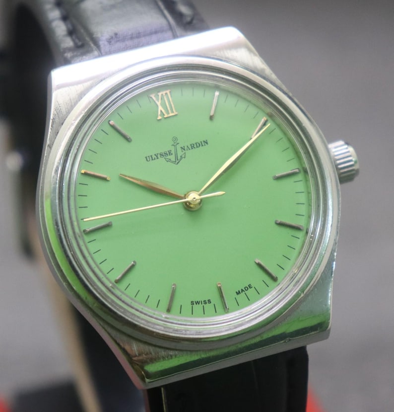 May include: A silver Ulysse Nardin wristwatch with a light green face. The watch has gold hands and hour markers, with the Roman numeral XII at the top. It features a black leather strap and the words "Ulysse Nardin" and "Swiss Made" are visible.