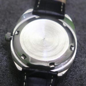 May include: Close-up of the back of a silver-toned wristwatch with a black leather band. The watch has a textured, circular back with a small, central indentation. The band is stitched and attached to the watch case.