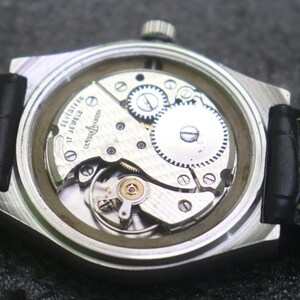 May include: A close-up view of a vintage Ulysse Nardin watch. The silver-tone case houses the intricate mechanical movement, visible through the clear back. The watch has a black leather strap. The watch face is not visible.