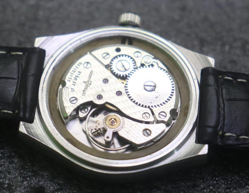 May include: A close-up view of a vintage Ulysse Nardin watch. The silver-tone case houses the intricate mechanical movement, visible through the clear back. The watch has a black leather strap. The watch face is not visible.