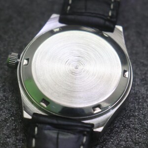 May include: A close-up of the back of a silver wristwatch with a black leather strap. The watch face is circular with a textured, brushed metal finish. The strap is stitched with white thread.