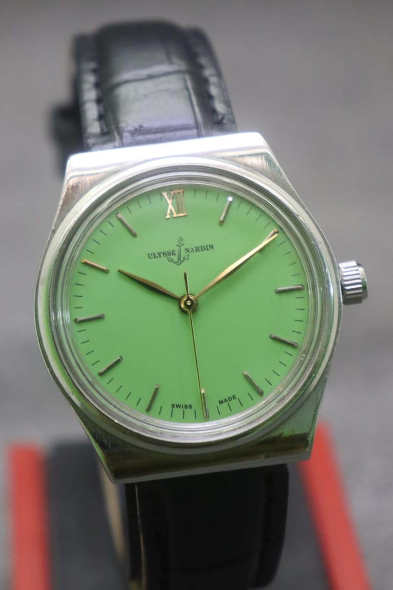 May include: A vintage Ulysse Nardin wristwatch with a bright green dial and gold hands. The watch has a silver-coloured case and a black leather strap. The dial features the brand name and the words "Swiss Made".