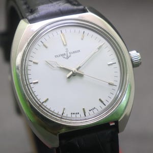 May include: A silver-toned Ulysse Nardin wristwatch with a white face and black leather band. The watch face features silver hands and markers, with the brand name and anchor logo. The watch is marked "Swiss Made".