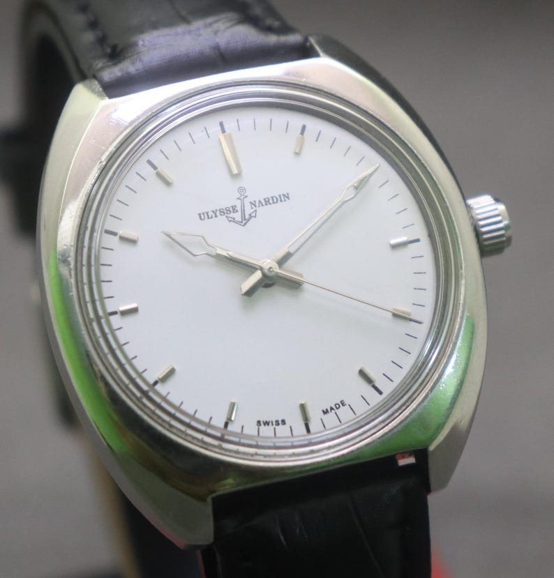 May include: A silver-toned Ulysse Nardin wristwatch with a white face and black leather band. The watch face features silver hands and markers, with the brand name and anchor logo. The watch is marked "Swiss Made".