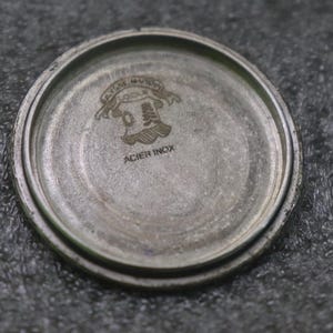 May include: A close-up of the inside of a silver watch case. The case has a circular shape with a raised edge. The text "ACIER INOX" is visible, along with an emblem.