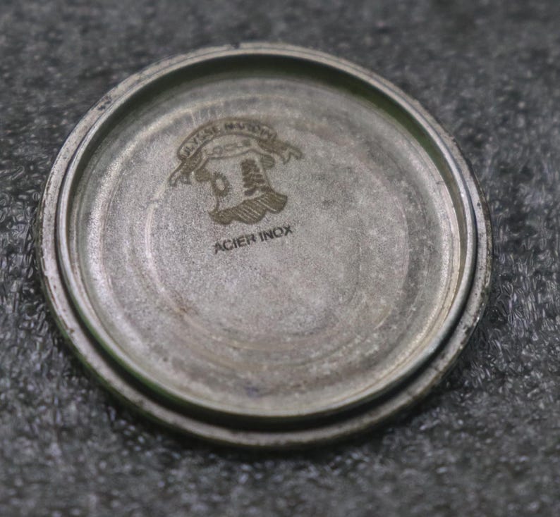 May include: A close-up of the inside of a silver watch case. The case has a circular shape with a raised edge. The text "ACIER INOX" is visible, along with an emblem.
