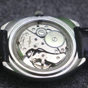 May include: A close-up of the back of a silver wristwatch with a black leather band. The watch face is clear, revealing the intricate internal mechanisms, including gears and jewels. The text "SWISS SEVENTEEN 17 JEWELS ULYSSE NARDIN" is visible.
