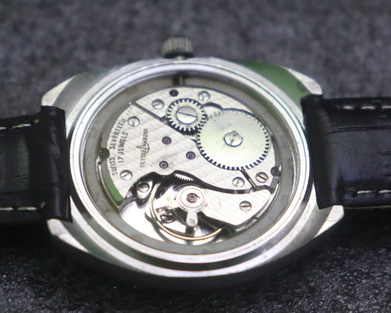 May include: A close-up of the back of a silver wristwatch with a black leather band. The watch face is clear, revealing the intricate internal mechanisms, including gears and jewels. The text "SWISS SEVENTEEN 17 JEWELS ULYSSE NARDIN" is visible.