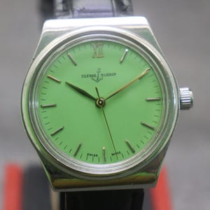 May include: A silver Ulysse Nardin wristwatch with a bright green face and a black leather strap. The watch has gold hands and markers, with the brand name and logo visible. The watch case is rectangular. The watch is Swiss made.