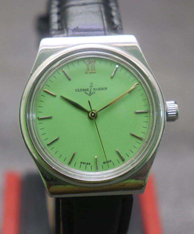 May include: A silver Ulysse Nardin wristwatch with a bright green face and a black leather strap. The watch has gold hands and markers, with the brand name and logo visible. The watch case is rectangular. The watch is Swiss made.