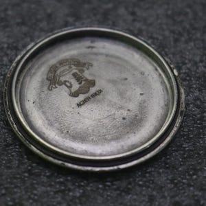 May include: Close-up of a watch back with a silver-coloured, circular design. The watch back has a textured, aged appearance. The centre features an engraved emblem with text above and below. The text "ACIER INOX" is visible.