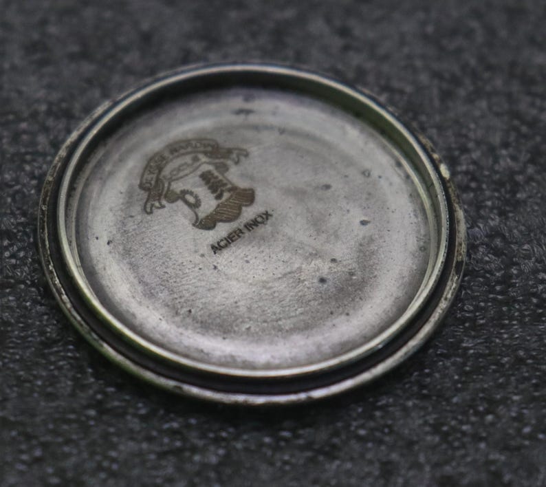 May include: Close-up of a watch back with a silver-coloured, circular design. The watch back has a textured, aged appearance. The centre features an engraved emblem with text above and below. The text "ACIER INOX" is visible.