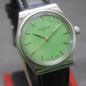 May include: A vintage Ulysse Nardin wristwatch with a bright green face and gold hands. The watch has a silver-coloured case and a black leather strap. The watch face displays the brand name and the word "SWISS MADE".