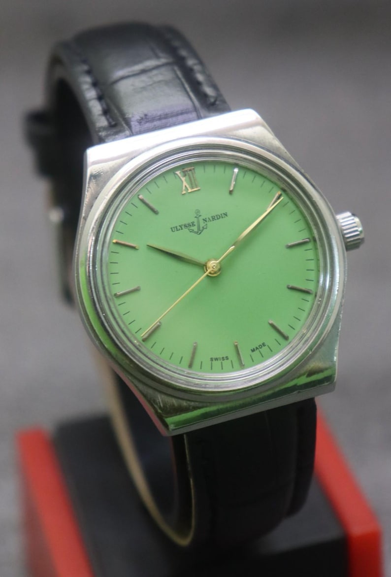 May include: A vintage Ulysse Nardin wristwatch with a bright green face and gold hands. The watch has a silver-coloured case and a black leather strap. The watch face displays the brand name and the word "SWISS MADE".