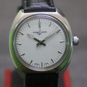 May include: A silver-toned Ulysse Nardin wristwatch with a white face and black hands. The watch features a black leather band and a silver crown. The watch face displays the brand name and the words "SWISS MADE".