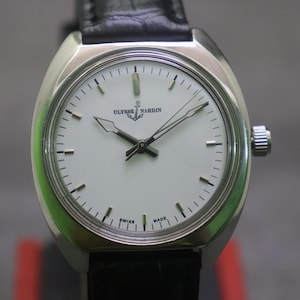 May include: A silver-toned Ulysse Nardin wristwatch with a white face and black hands. The watch has a black leather band and a silver crown. The watch face displays the brand name and the words "SWISS MADE".