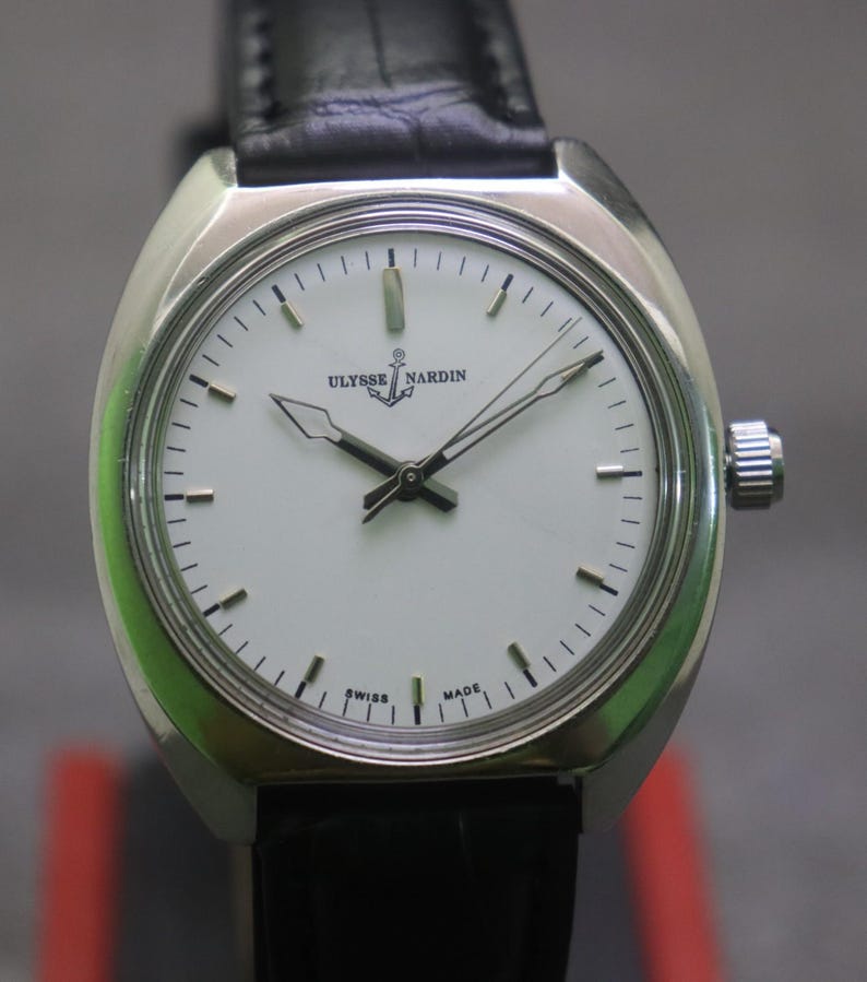 May include: A silver-toned Ulysse Nardin wristwatch with a white face and black hands. The watch has a black leather band and a silver crown. The watch face displays the brand name and the words "SWISS MADE".
