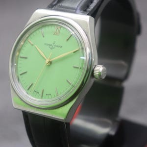 May include: A vintage Ulysse Nardin wristwatch with a light green dial and gold hands. The watch has a silver-tone case and a black leather strap. The dial features the brand name and the words "Swiss Made".