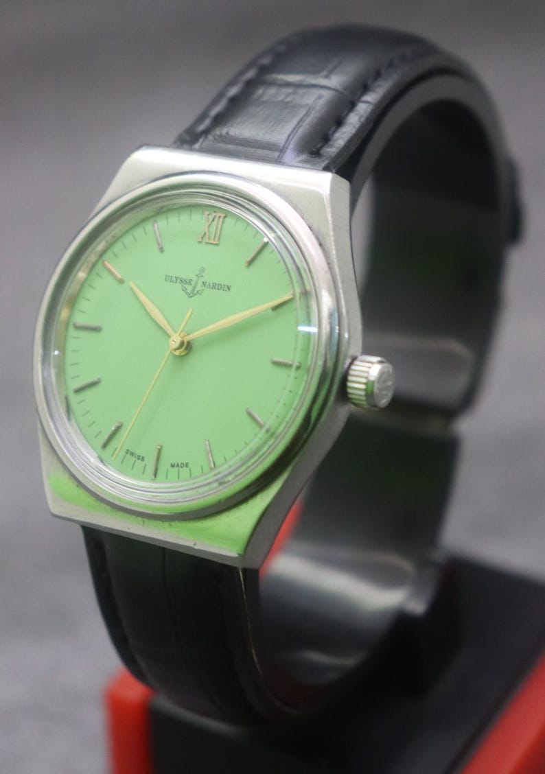 May include: A vintage Ulysse Nardin wristwatch with a light green dial and gold hands. The watch has a silver-tone case and a black leather strap. The dial features the brand name and the words "Swiss Made".