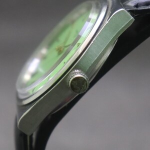 May include: Close-up of a vintage wristwatch with a light green face and silver-coloured casing. The watch has a black leather strap and a textured crown. The watch face has a gold-coloured design.