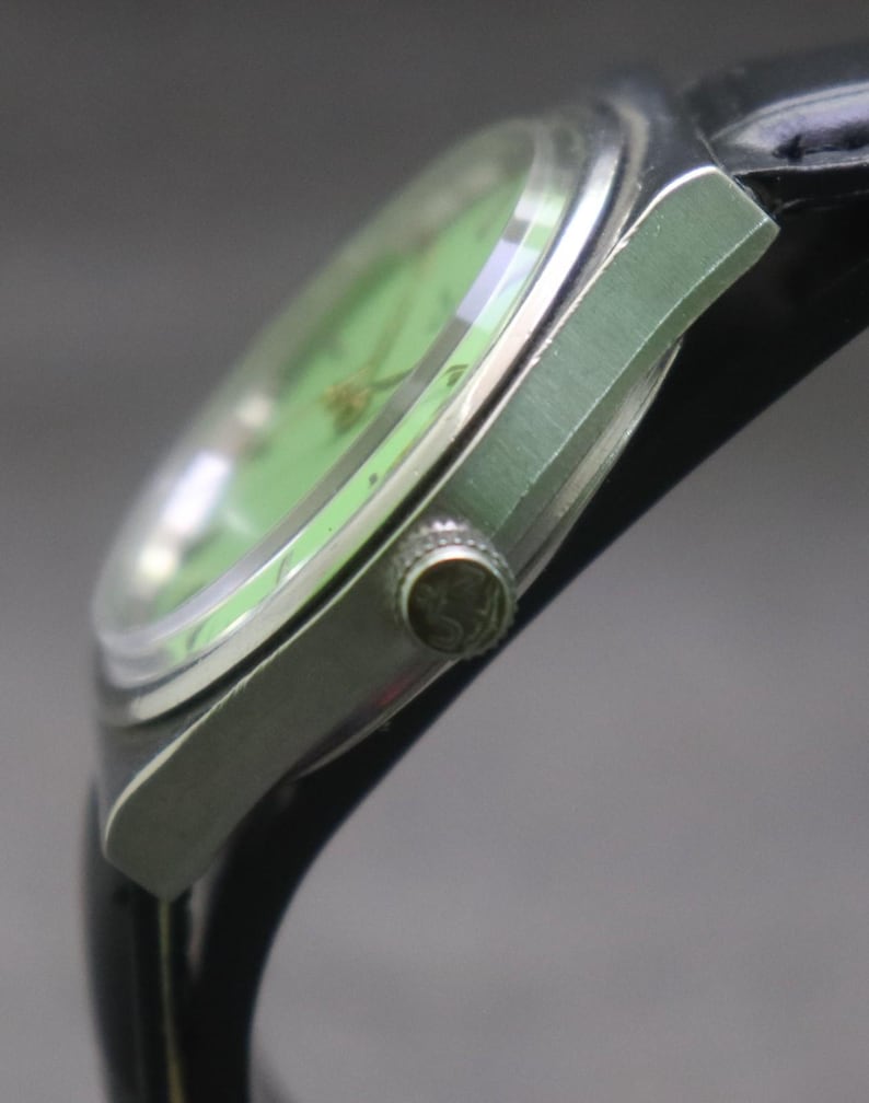 May include: Close-up of a vintage wristwatch with a light green face and silver-coloured casing. The watch has a black leather strap and a textured crown. The watch face has a gold-coloured design.