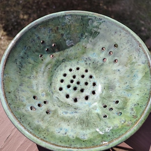 May include: A handmade ceramic colander with a speckled green glaze. The bowl has a series of drainage holes and a slightly raised rim. The color palette includes shades of green and blue, creating a unique kitchenware piece.