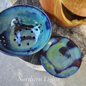 May include: A ceramic berry bowl with a matching saucer, featuring a glossy glaze in swirling shades of deep blue, teal, and lime green. The bowl has drainage holes at the bottom. The text Northern Lights is printed at the bottom.
