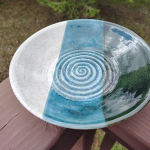 May include: A handmade ceramic bowl with a spiral design in the center. The bowl is divided into three sections: white speckled, teal, and dark teal. The bowl is sitting on a wooden surface.
